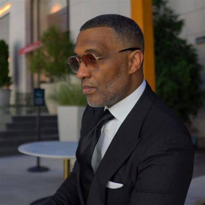 Kevin Samuels is wearing a black turtle neck, grey suit and a spec in the picture.